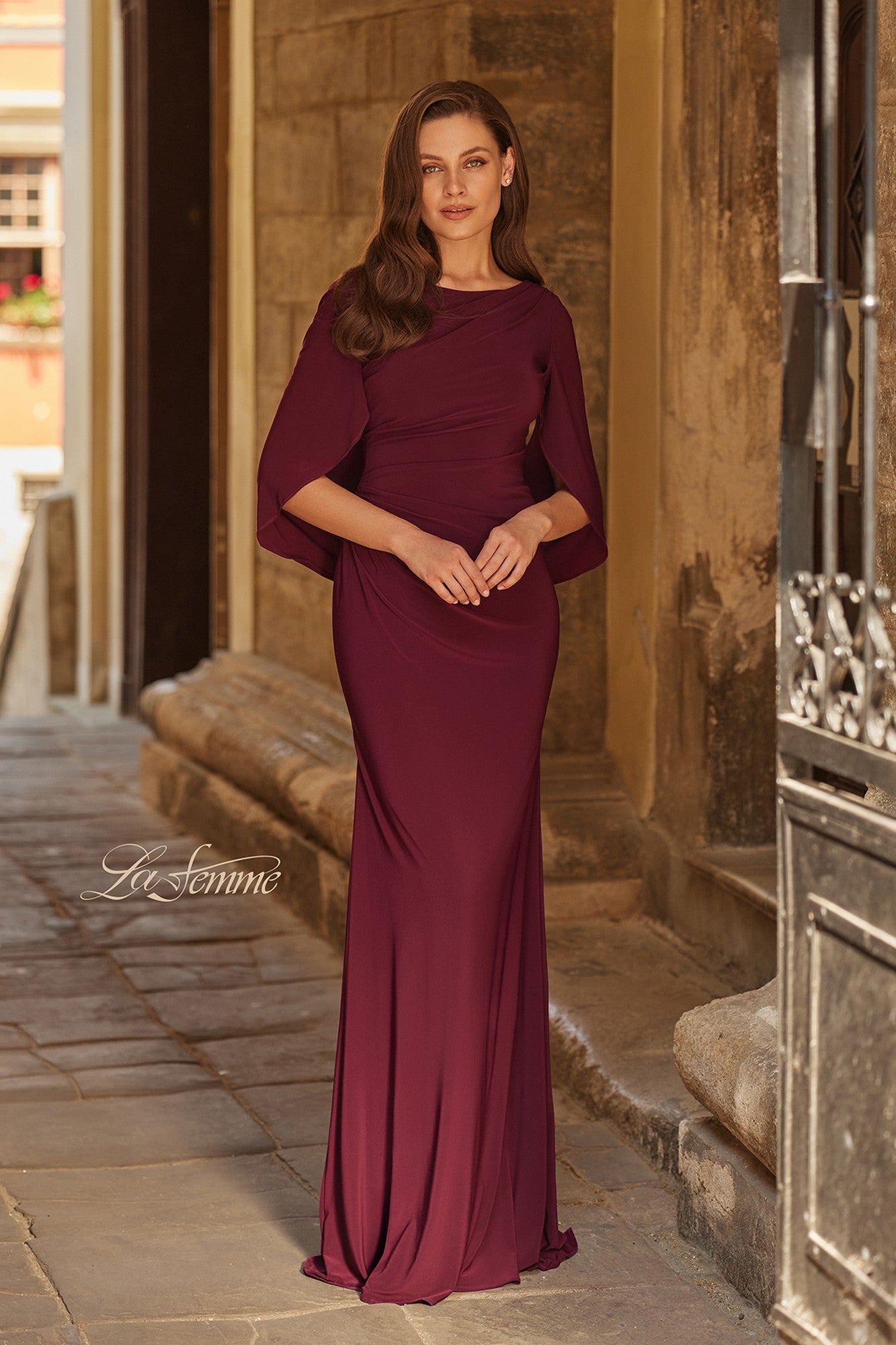A woman stands in a stone corridor wearing a La Femme 33148 long formal dress in dark berry, featuring elegant cape sleeves.
